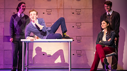 Four students on stage for a play with one male student laying playfully on a table calling out with his hand near mouth