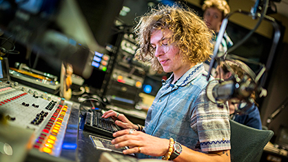 A male student with shaggy hair sits at the WNUR radio console panel pressing orange and red blinking buttons to control audio levels of the show