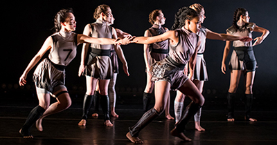 Six female dancers in gray outfits with shorts face stage right while two clasp hands