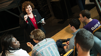 Professor Cindy Gold speaks while smiling in front of dark room with students listening intently in foreground