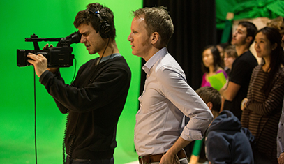 With green screen in background, professor observes and directs student in headset looking through video camera