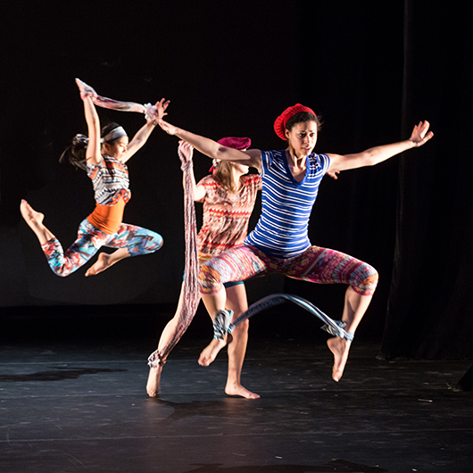 On a dark stage three female dancers in midair jumps wearing colorful outfits of red, blue and orange and orange