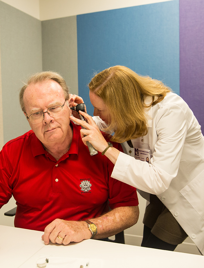 Female practitioner in white coat uses an otoscope to examine the inner ear of an older man in red shirt